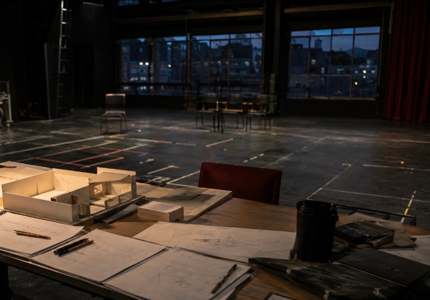 A white card model of a set on a drawing table, surrounded by technical drawings, pencils and a paint tin, in a rehearsal room whose windows show a city at dusk.