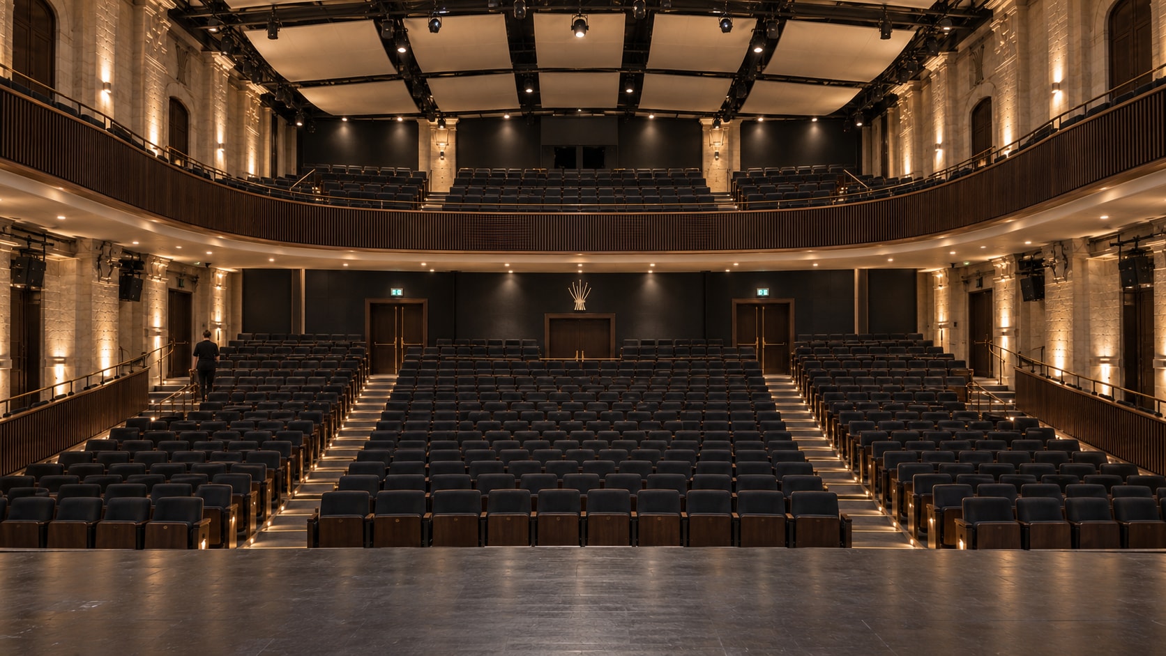 The empty house of the Foundation’s theatre: raked seating in dark wood rising toward a shallow balcony, warm light washing the side walls, the room waiting.