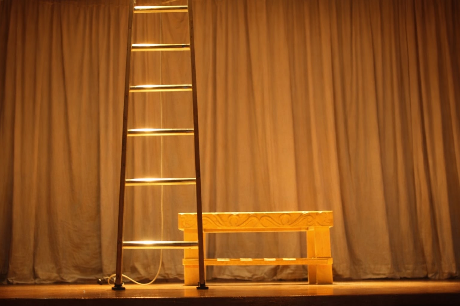 An empty stage: a ladder and a carved bench against a warm curtain.