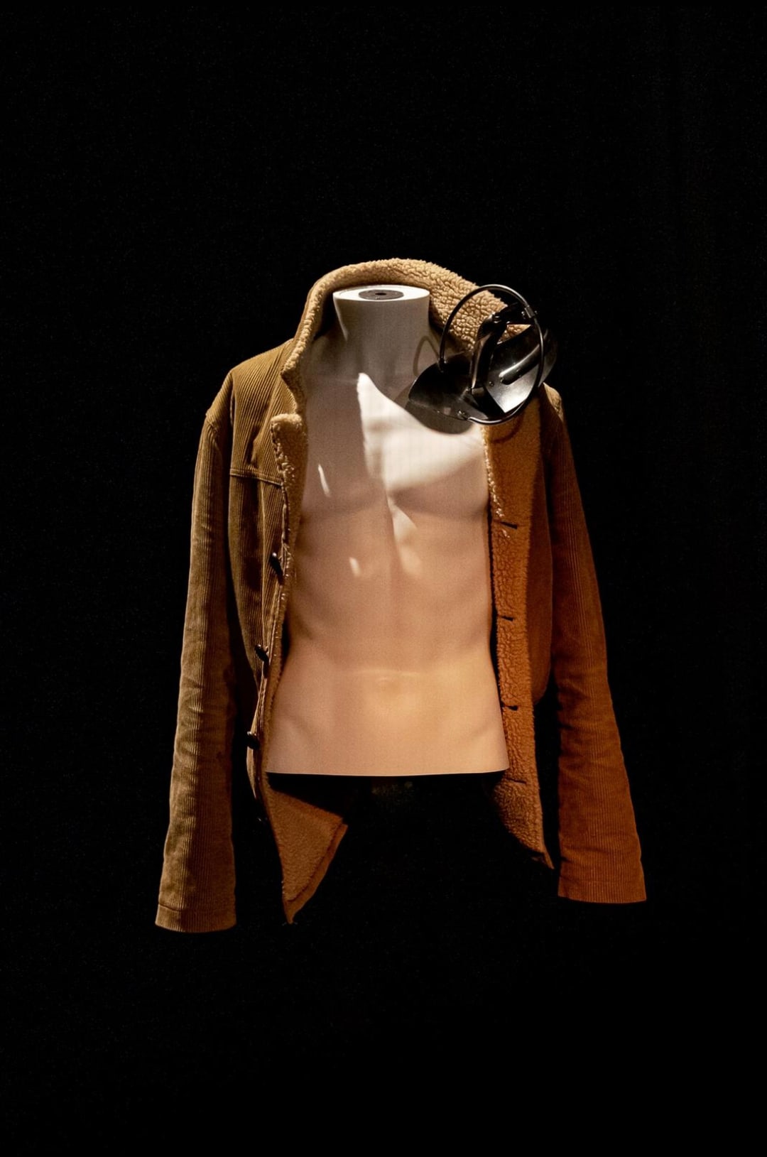 A corduroy jacket with a shearling lining, worn open on a mannequin torso and lit against black, a stage lamp resting against its shoulder.