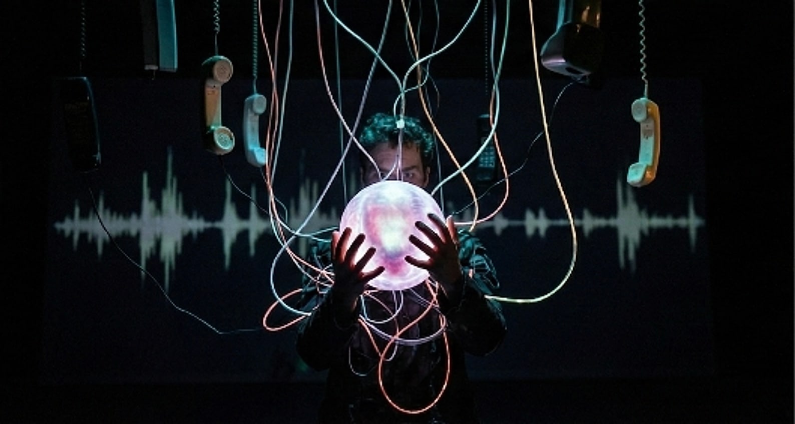 A performer cupping a glowing sphere in both hands, surrounded by telephone handsets hanging on tangled cables, an audio waveform projected across the wall behind.